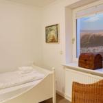 Ein Schlafzimmer mit Bett, Fenster und Kiste. Draußen Sanddünen und Holzweg.