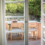 Durchgehende Glastüren führen auf eine Terrasse mit Holztisch und Stühlen. Hintergrund: grüne Hecke und Steinmauer.