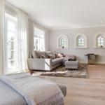 Wohnzimmer mit Sofa, Teppich und drei Bogenfenstern. Holzboden und Kamin sichtbar.