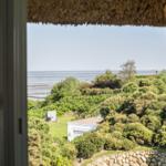 Durch das Fenster blickt man auf einen Garten mit Bäumen und das Meer.
