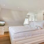 Schlafzimmer mit weißem Bett, TV und Holzboden. Dachfenster und Spiegel sorgen für Licht.