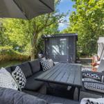 Terrasse mit Sitzgruppe, Tisch und Sonnenschutz. Hintergrund: Gartenhaus und Bäume.