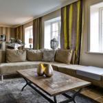 Ein Wohnzimmer mit Sofa, Tisch und Fenstern. Vorhänge und Wandstreifen sind sichtbar.