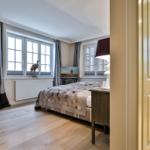 Schlafzimmer mit Bett, Fenstern und Holzboden. Tür führt in Bad.