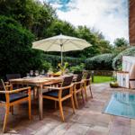 Terrasse mit Tisch, Stühlen und Sonnenschirm. Garten mit Rasen und Hecken. Glasplatte neben Hauswand.