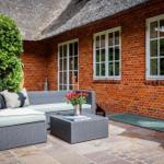 Terrasse mit Rattan-Sofa, Tisch und Blumen vor rotem Backsteinhaus mit Strohdach.