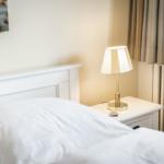 Schlafzimmer mit weißem Bett, Nachttisch und Lampe. Fenster mit Vorhängen.