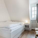 Schlafzimmer mit weißem Bett, Nachttisch und Fenster.