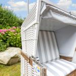 Weiße Strandkorb mit Streifenpolster im Garten, umgeben von Grün und Blumen.