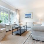 Wohnzimmer mit hellen Sofas, Glas-Tisch und großen Fenstern mit Blick auf Grün.