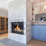 Kamin mit Flammen, Holzboden, Küchenzeile und hängende Lampen.
