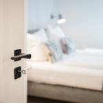 Schlafzimmer mit Bett und Nachttischlampe. Tür mit schwarzer Klinke ist geöffnet.