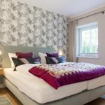 Schlafzimmer mit Bett, Kopfteil, Wandtapete und zwei Fenstern.