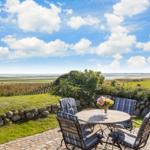 Terrasse mit Tisch und Stühlen, Blick auf Wiesen und Meer unter blauem Himmel.