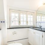 Badezimmer mit Fenster, Waschbecken und Toilette. Weiße Fliesen und dunkler Arbeitsplatz.