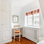Schlafzimmer mit weißem Schrank, Schreibtisch und Fenster mit buntem Rollladen.