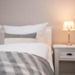 Ein Bett mit weißen Kissen und einer grau-weißen Decke neben einem Nachttisch mit Lampe.