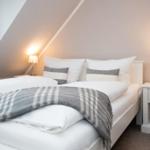 Schlafzimmer mit Dachschräge, Bett mit weißen Laken und grauer Decke, zwei Nachttische mit Lampen.