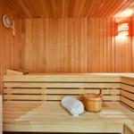 Sauna mit Holzbank, weißem Handtuch und Eimer. Wanduhr und Wandlampe sichtbar.