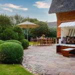 Terrasse mit Strandkorb, Tisch und Stühlen, umgeben von grünen Büschen und einem Steinweg.