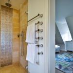 Dusche mit Glaswand und Handbrause. Badetuchhalter mit weißen Handtüchern. Blick in Schlafzimmer mit Teppich.