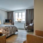 Schlafzimmer mit Bett, TV und Sessel. Holzboden und Fenster mit Vorhängen.