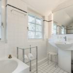 Badezimmer mit Badewanne, Waschbecken und zwei Fenstern mit Jalousien.