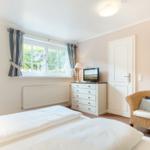 Schlafzimmer mit Bett, Schrank, Fernseher und Sessel. Fenster mit Blick auf Grün.
