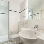 Badezimmer mit Dusche, Waschbecken und Toilette. Weiße Fliesen und Spiegel.