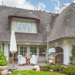 House with thatched roof, terrace with table and chairs, garden with shrubs and stones.