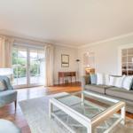 Wohnzimmer mit Sofa, Sessel und Glas-Tisch. Holzboden und Fenster zur Terrasse.