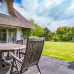 Haus mit Strohdach, Terrasse mit Tisch und Stühlen, grüner Garten im Hintergrund.