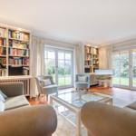 Wohnzimmer mit Bücherregalen, Sesseln und Glas-Tisch. Durchgänge zu Garten.