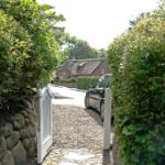 A white gate leads through green hedges to a courtyard with a car and a house in the background.