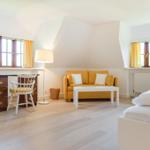 Zimmer mit gelbem Sofa, Schreibtisch und Bett. Holzboden und große Fenster.