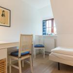 Schlafzimmer mit Schreibtisch, Stühlen und Bett. Fenster mit blauen Vorhängen.