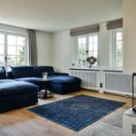 A living room with blue sofa, wooden floor, and windows with curtains.