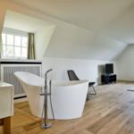 Attic room with freestanding bathtub, wooden floor, and windows.