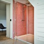Shower with red tiles and glass enclosure. Radiator on the wall. Door leads to a room with a chair.