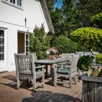 Bright terrace with wooden table, chairs, and flower arrangement. White house wall and green plants in background.