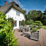 White house with thatched roof, terrace with table and chairs, landscaped garden with trees.