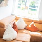 Ein braunes Sofa mit Kissen in Orange und Weiß vor einem Fenster.