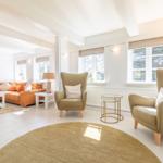 Wohnzimmer mit Sofa, zwei Sesseln und großen Fenstern.