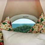 Rundes Fenster mit Blick auf Meer und Bäume, zwei Kissen mit Blumenmuster vorne