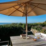 Terrasse mit Tisch, Sonnenschirm und Meerblick. Blumenbeete und ein traditionelles Haus sind sichtbar.
