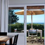 Terrasse mit Tisch, Stühlen und Sonnenschirm. Blick auf Meer und Bäume.