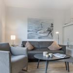 Wohnzimmer mit grauem Sofa, Sessel und runden Tisch. Wandbild und Fenster sichtbar.