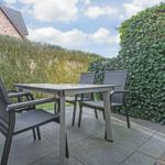 Terrasse mit Tisch und vier Stühlen, umgeben von grünen Hecken und einem Haus.