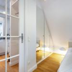 Schlafzimmer mit Dachschrägen, Bett, Lampe und Glaswand. Tür führt in Küche mit Fenster.