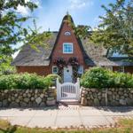 Red brick house with thatched roof and white door. Garden with stone wall and flowers.